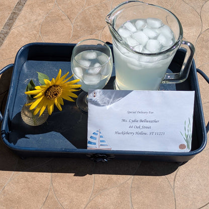Envelope addressed to Lydia Bellweather from The Vanishing Compass 12 month cozy mystery by mail, on a blue metal tray with a Sunflower and glass and pitcher of lemonade with ice - all on a stone-top patio table